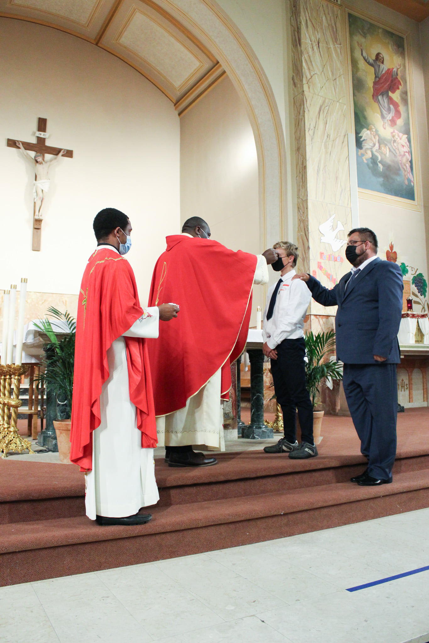 Fr. Simon confirming confirmation candidate with Fr. Ben to his right and the sponsor of candidate behind candidate.